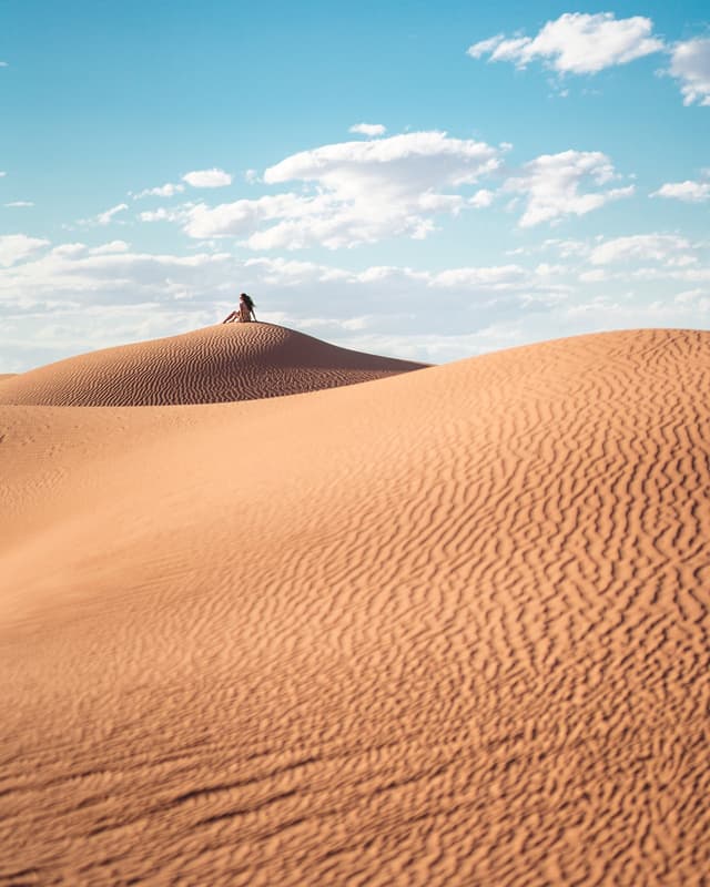 Person sitting on enormous sand dunes with fluffy clouds in a blue sky behind them